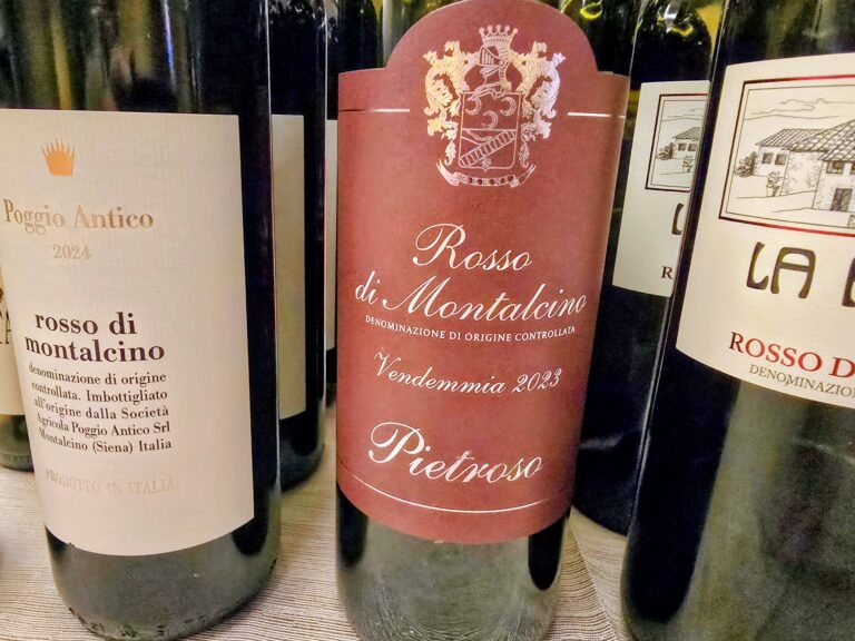 Bottiglia di Pietroso Rosso di Montalcino 2023 con etichetta bordeaux su sfondo di altre bottiglie in degustazione