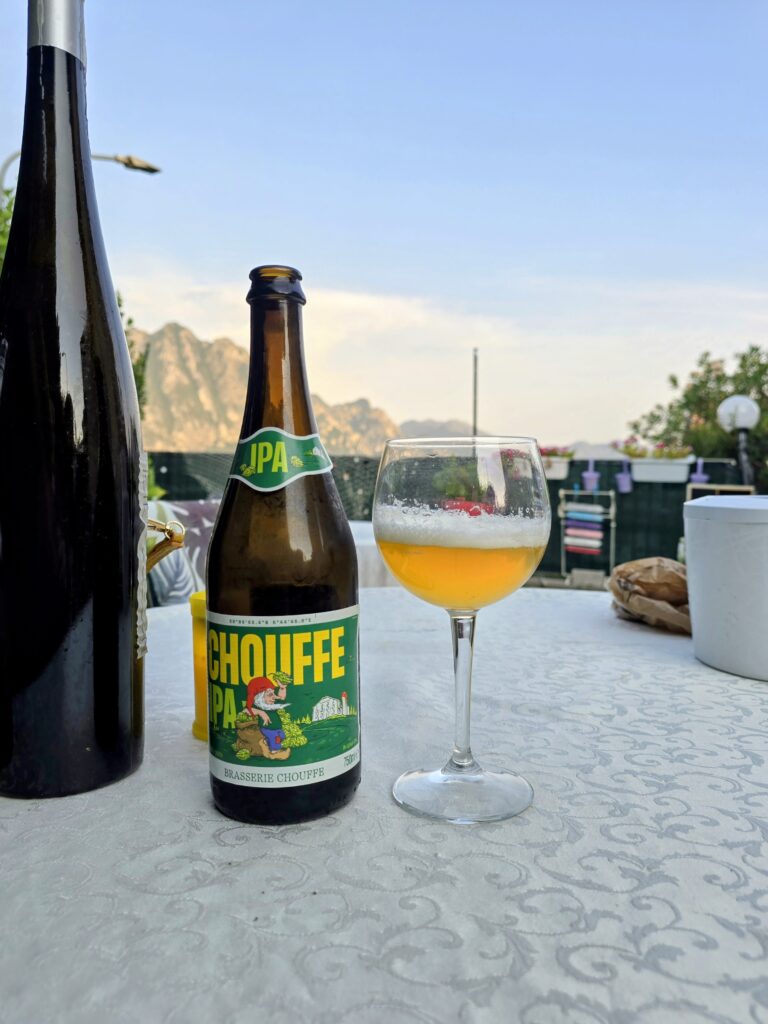 Bicchiere di Chouffe IPA dorata con schiuma bianca persistente accanto alla bottiglia da 750 ml, su terrazza al tramonto