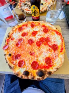 Pizza bassa con salsiccia piccante a fette su tavolo in legno, servita con Leffe Rouge, al ristorante Trentapassi di Riva di Solto