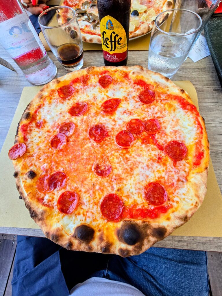 Pizza bassa con salsiccia piccante a fette su tavolo in legno, servita con Leffe Rouge, al ristorante Trentapassi di Riva di Solto