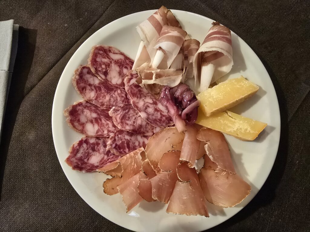 Tagliere di salumi visto dall'alto: fette di salame a grana grossa, pancetta arrotolata, lonza stagionata e due tranci di formaggio su un piatto bianco